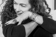 Monochrome photo of a woman with tight curls, resting her cheek on folded arms that showcase studded rings, bracelets, and earings.